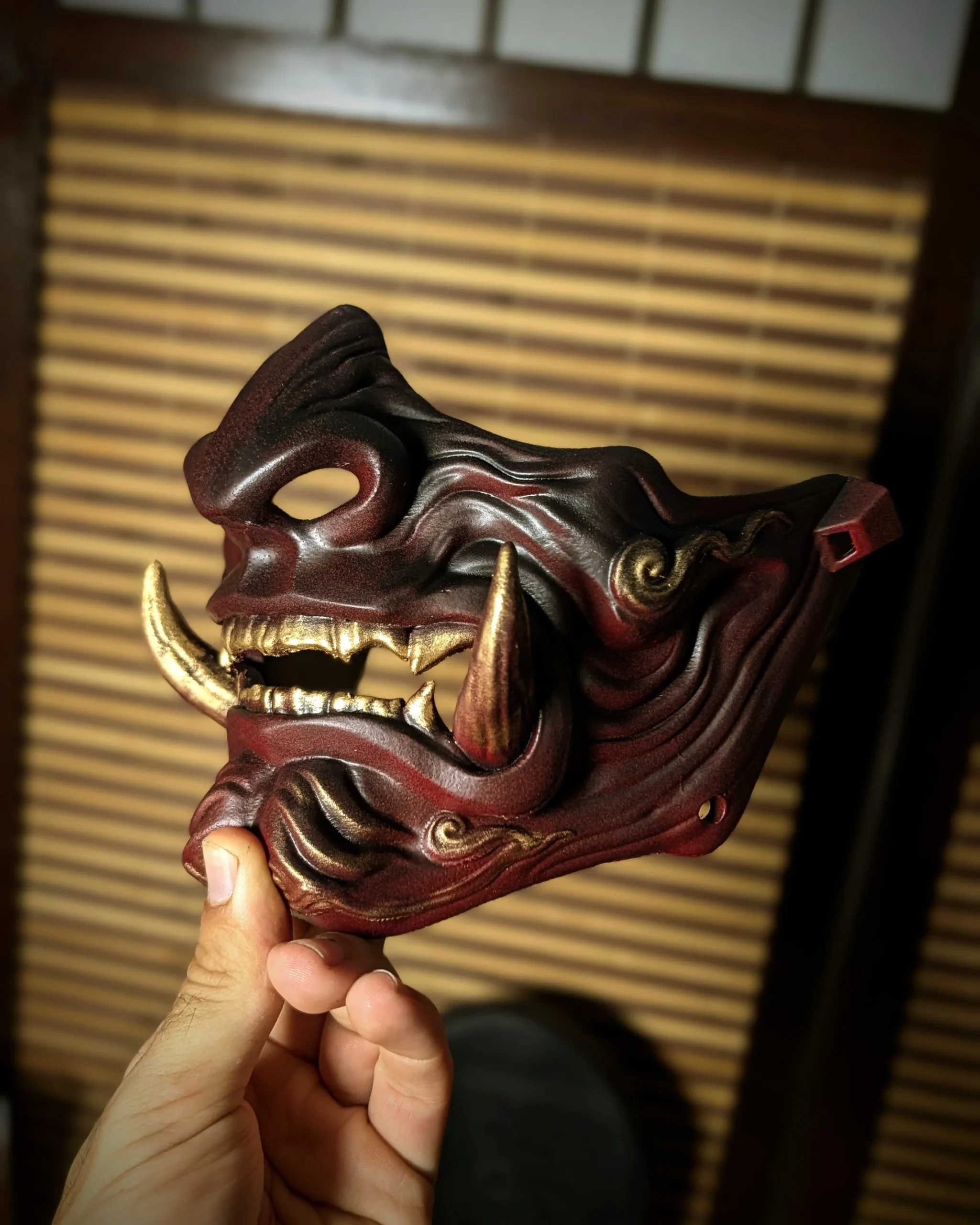 Demi-masque Mempo Oni rouge à dents dorées, tenu en main