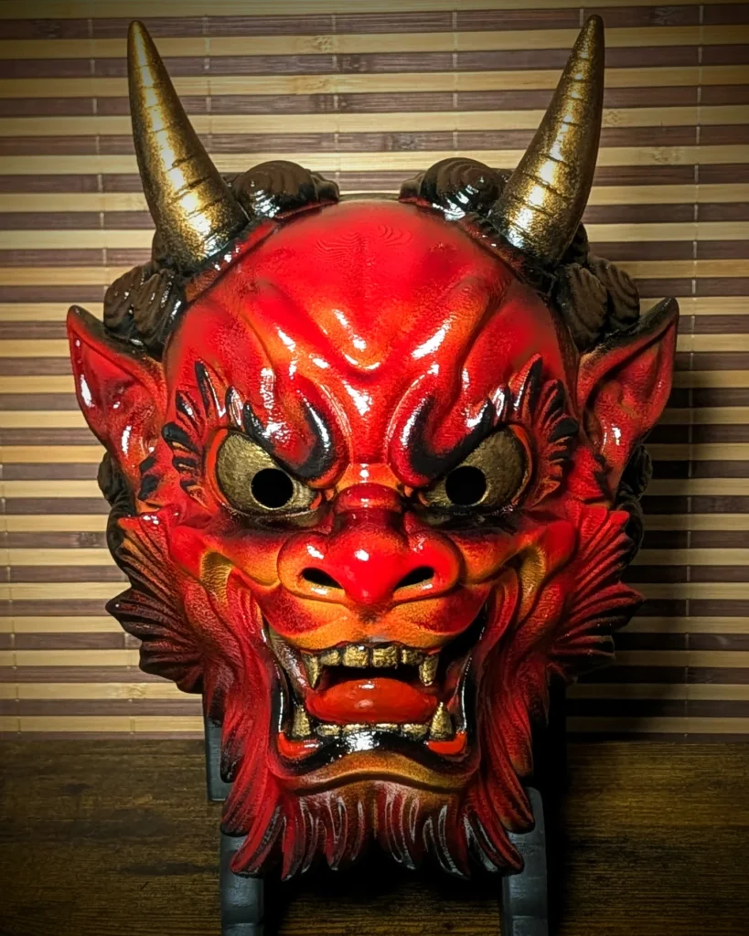 Máscara Oni roja artesanal con cuernos dorados, expuesta de frente sobre un soporte